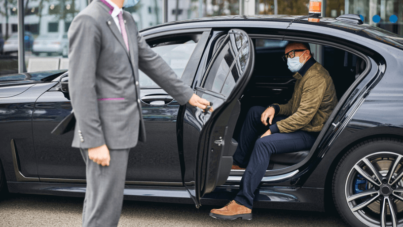 Chauffeur opening the car door for a Caucasian male passenger in a disposable face mask