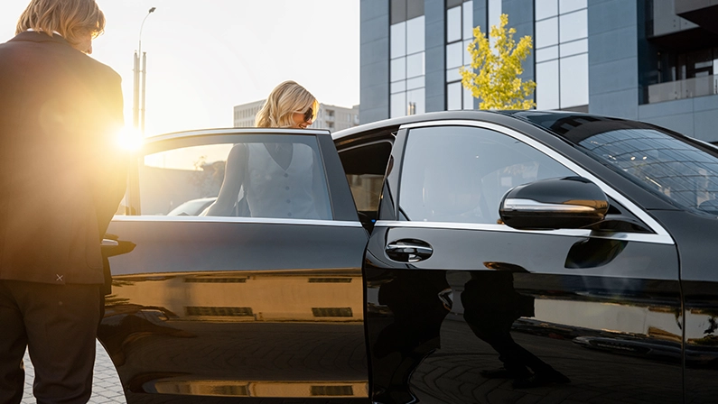 Business man helps business lady to get in the car, opening vehicle door near office building on sunset. Concept of business lifestyle and transportation